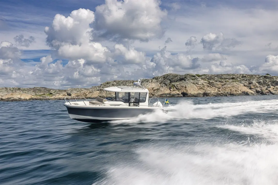 A white motorboat speeds across calm waters, leaving a wake, with rocky shores and fluffy clouds in the background.